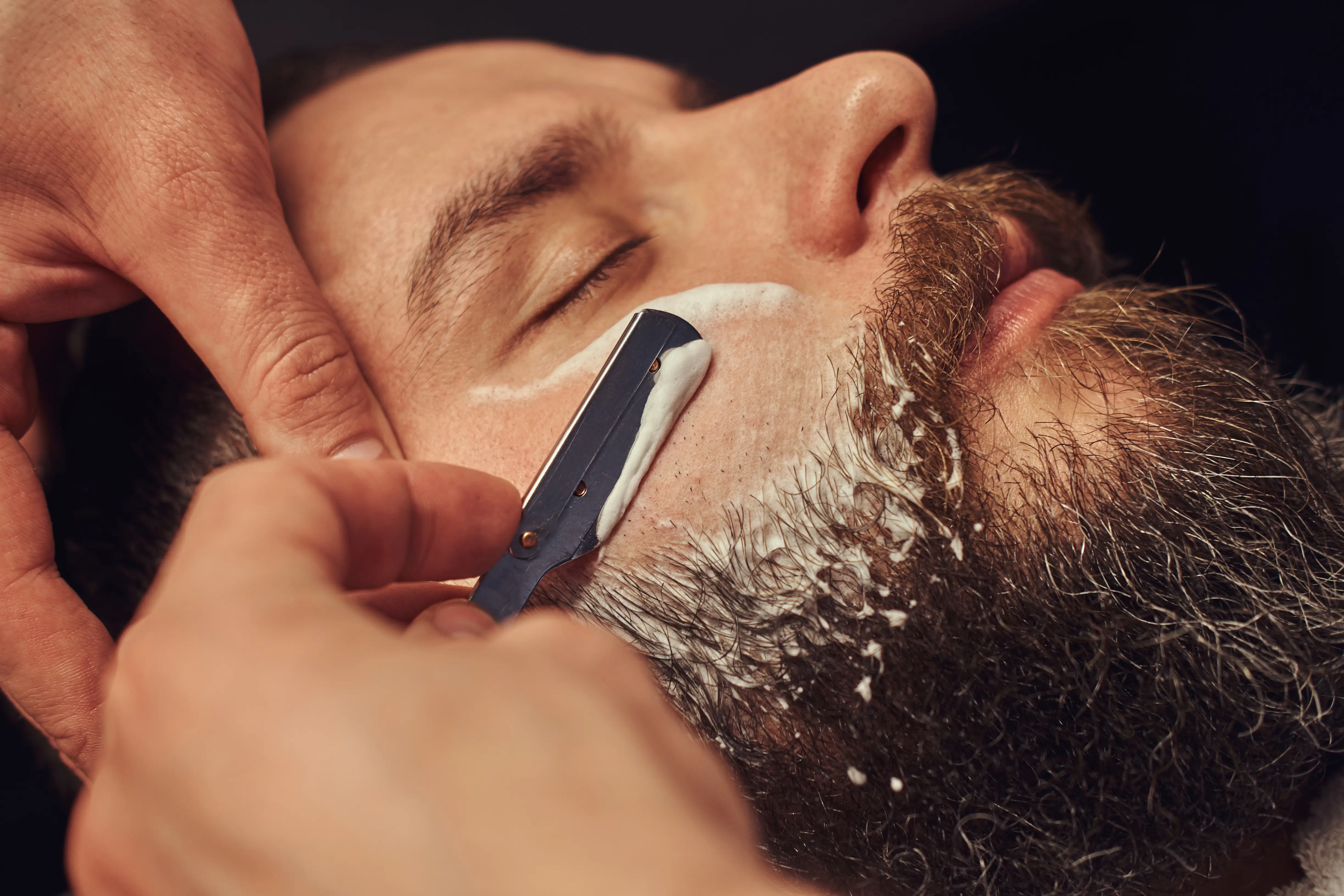 A barber drawing a straight razor through lather along a client's jaw.